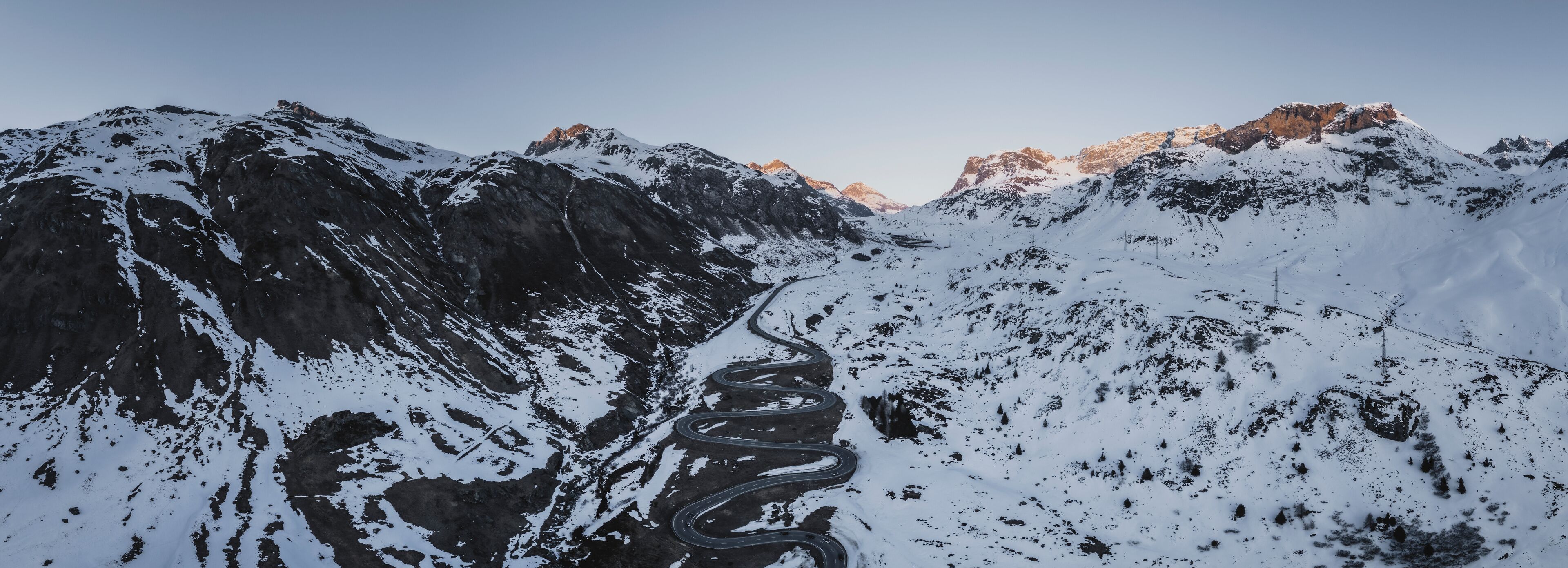 A winding mountain road through a snow-covered landscape with rocky outcrops and distant peaks.