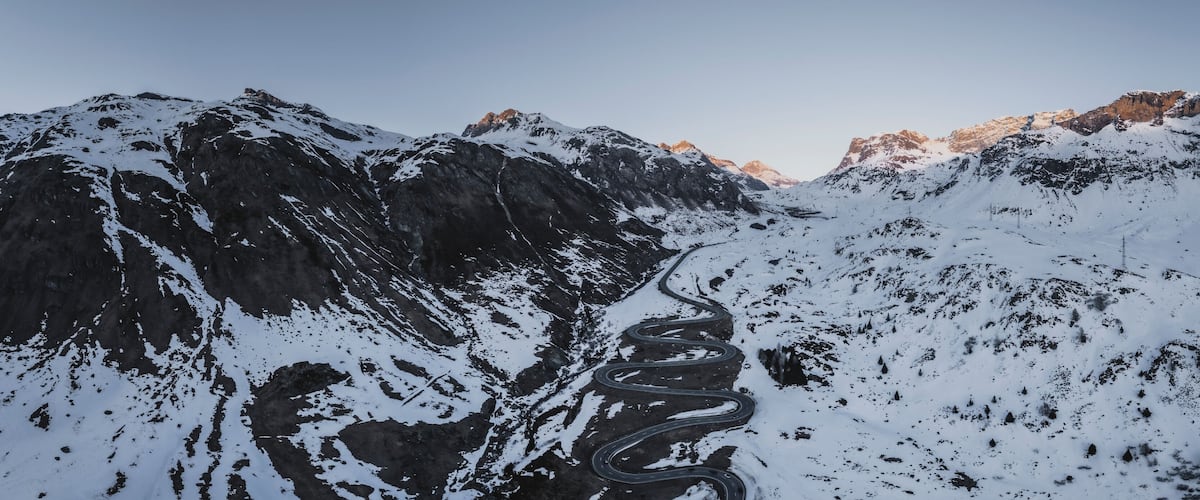 A winding mountain road through a snow-covered landscape with rocky outcrops and distant peaks.