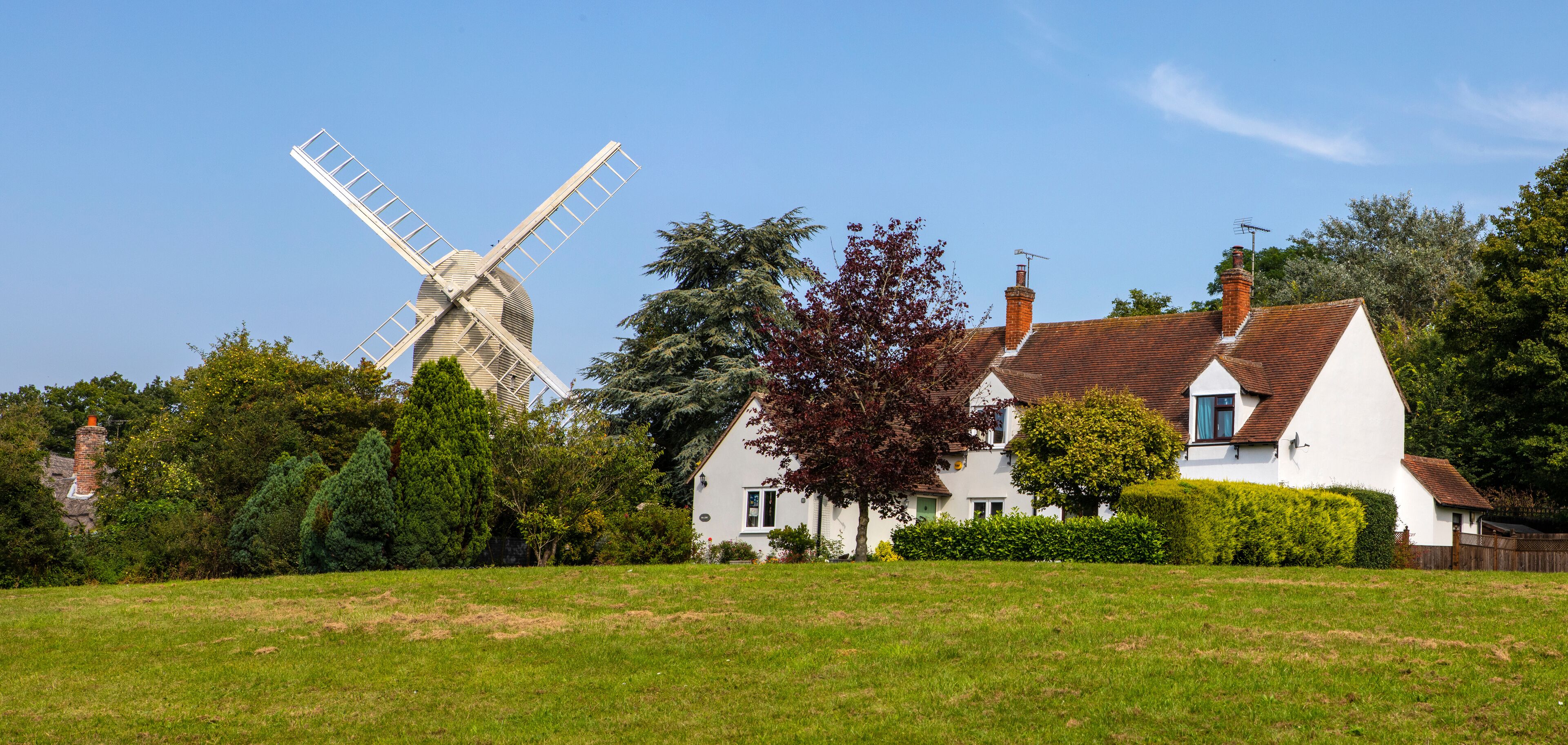 A white windmill, a house with a red roof, and lush green trees in a rural setting.