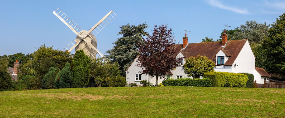 A white windmill, a house with a red roof, and lush green trees in a rural setting.