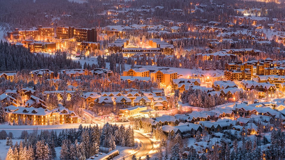 A snow-covered resort town at dusk with illuminated buildings and a winding road.