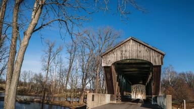 A covered bridge with a wooden structure and a metal roof, surrounded by leafless trees and a body of water.