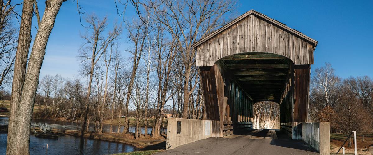 A covered bridge with a wooden structure and a metal roof, surrounded by leafless trees and a body of water.