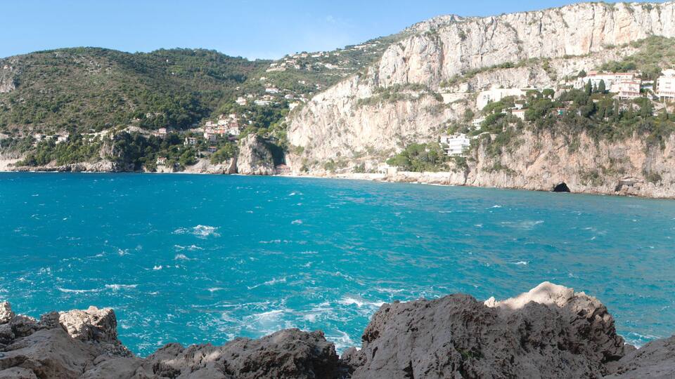 A coastal landscape with a clear blue sea, rocky cliffs, and buildings on the hillside.
