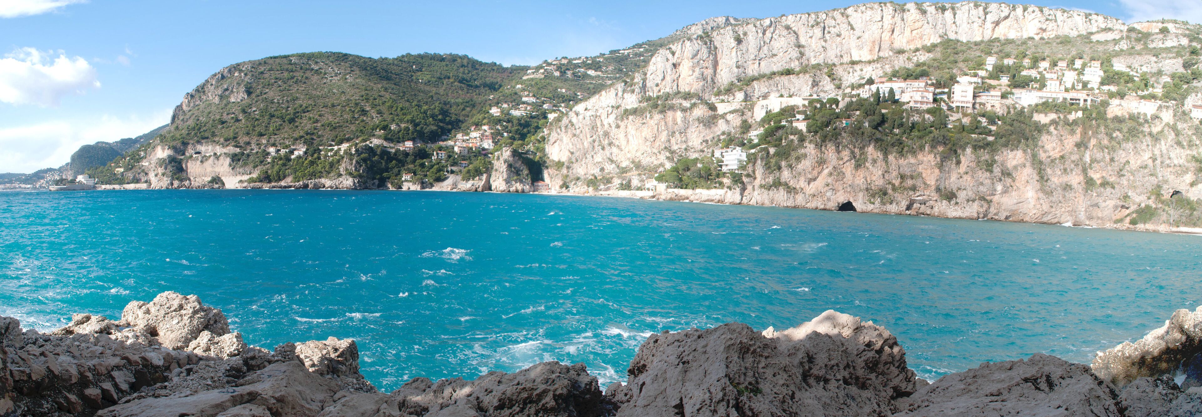 A coastal landscape with a clear blue sea, rocky cliffs, and buildings on the hillside.