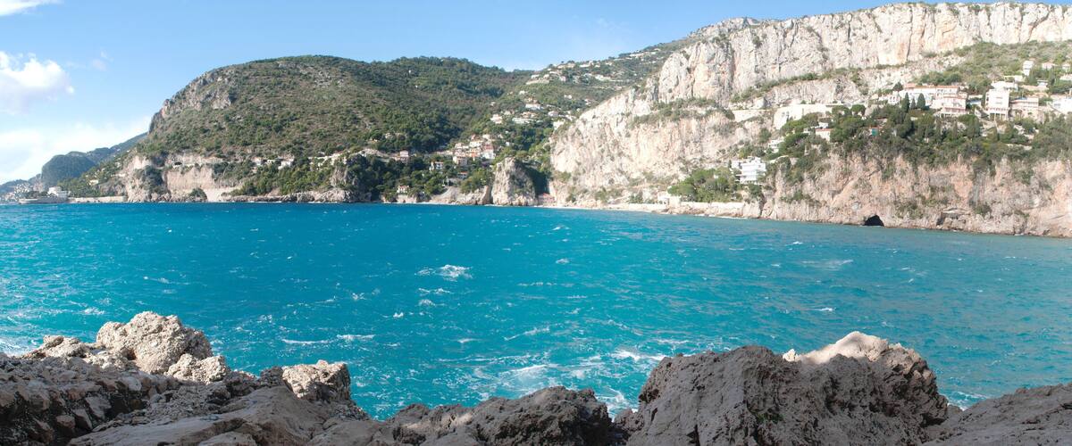 A coastal landscape with a clear blue sea, rocky cliffs, and buildings on the hillside.