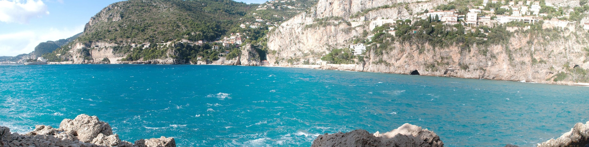 A coastal landscape with a clear blue sea, rocky cliffs, and buildings on the hillside.