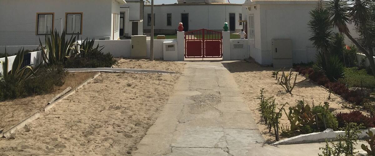 A lighthouse with a red top, a white building with a red gate, and a sandy path leading to it.