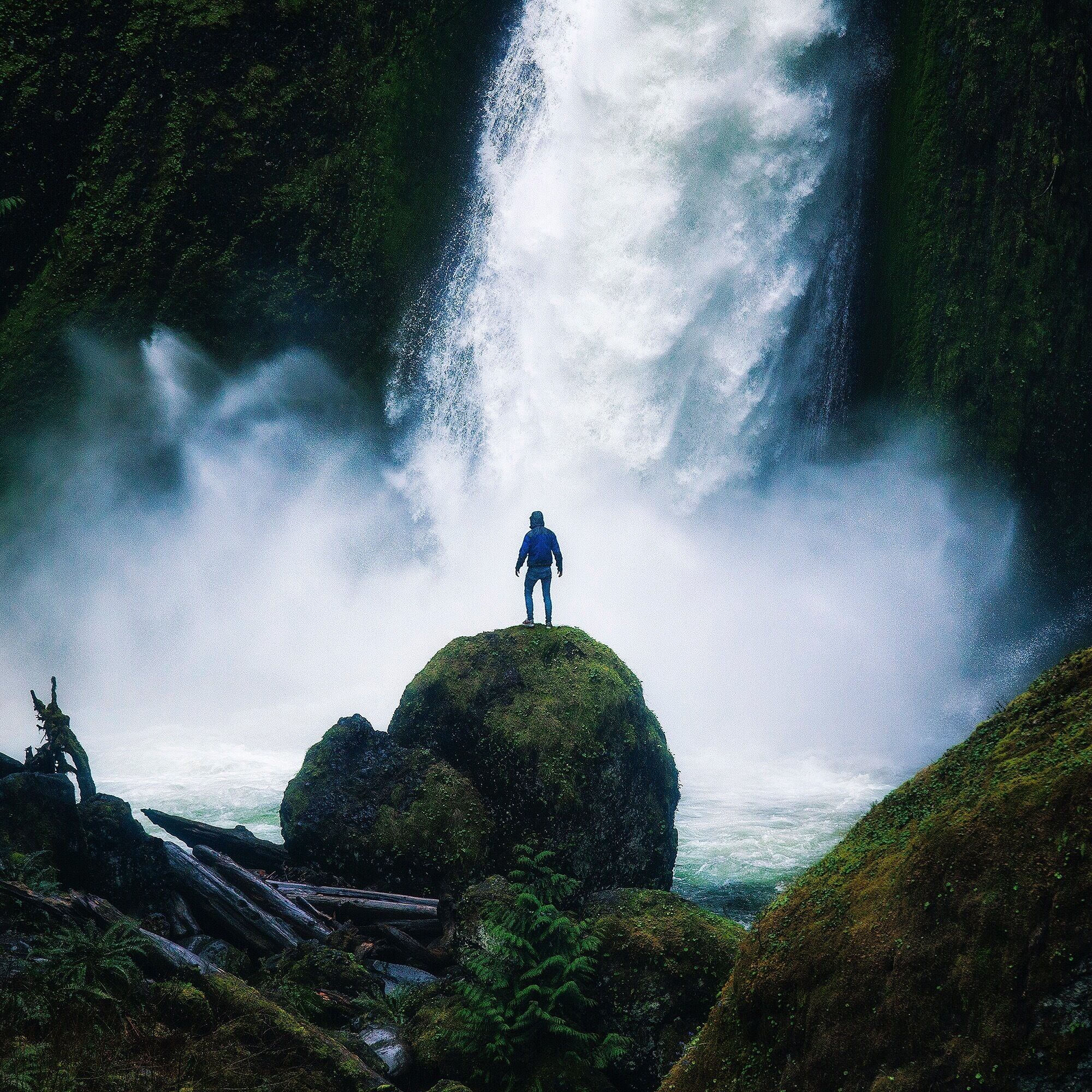 A person standing on a moss-covered rock with a waterfall in the background.