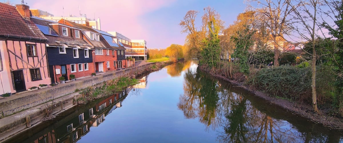 A calm river reflecting buildings and trees, with a mix of traditional and modern architecture.