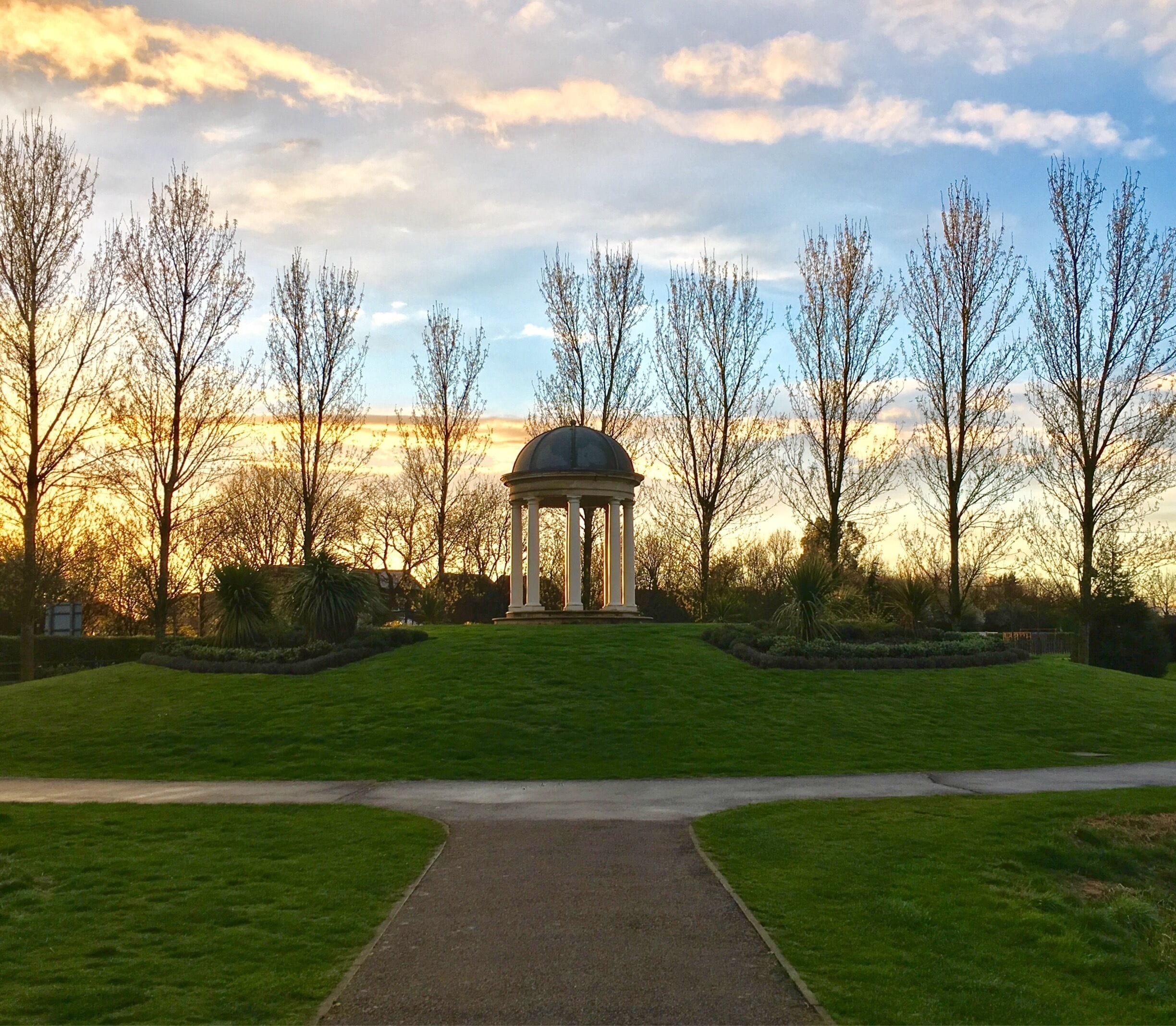 A gazebo in a park with a pathway leading to it, surrounded by trees and a sunset in the background.