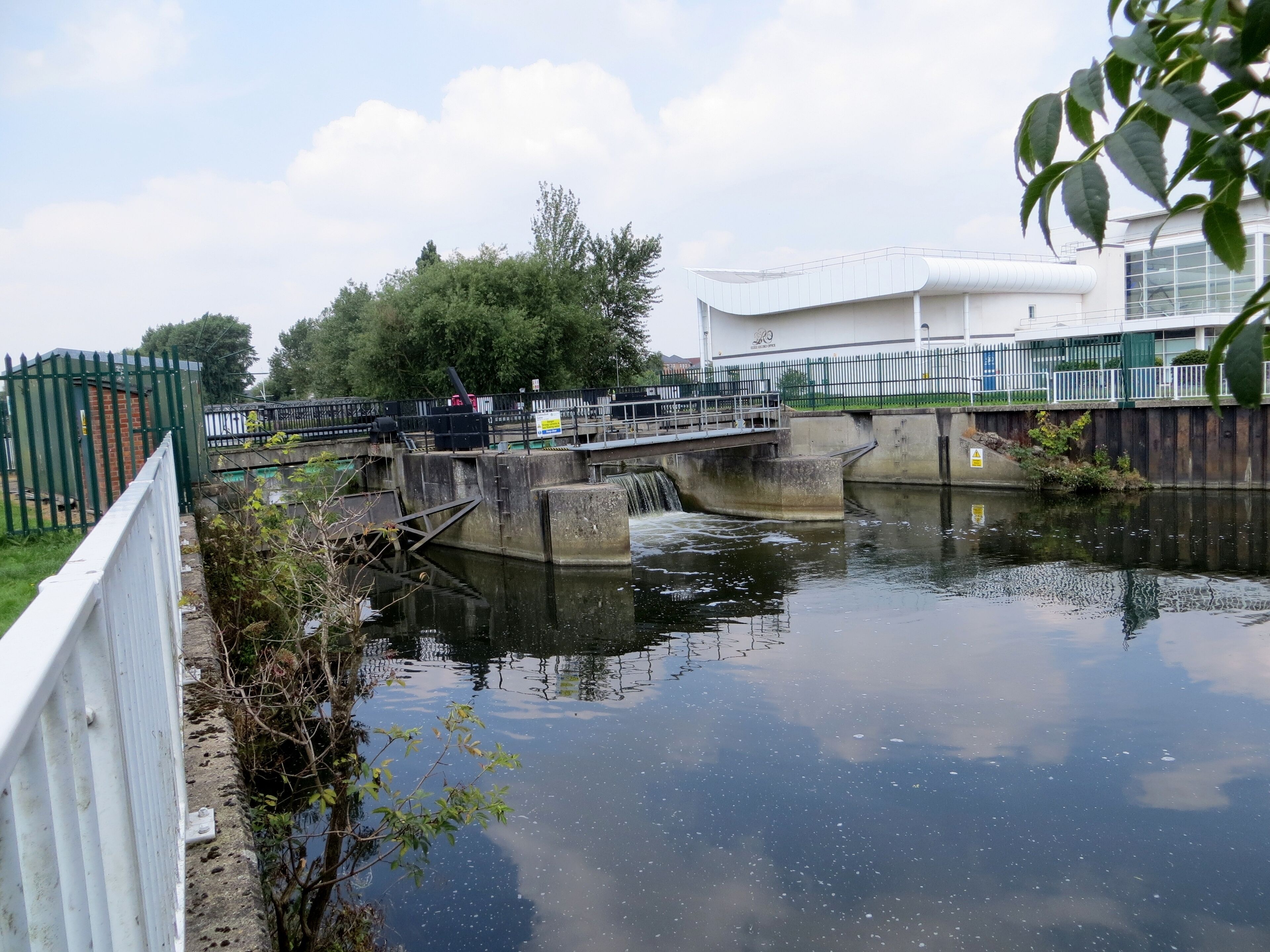 A lock on a canal with a bridge crossing over it, surrounded by greenery and a building in the background.