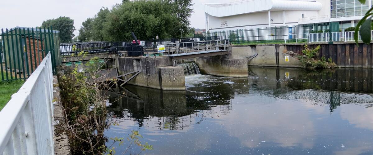 A lock on a canal with a bridge crossing over it, surrounded by greenery and a building in the background.
