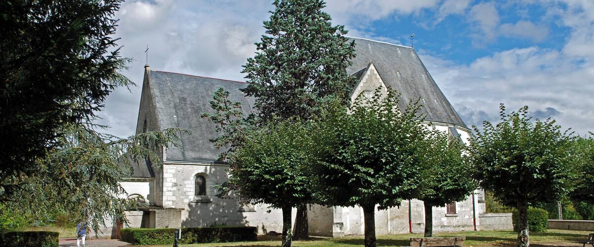 A historic stone building with a steep roof, surrounded by trees and a paved path.