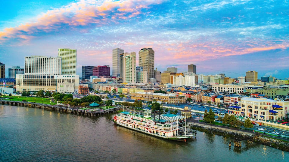 A river with a steamboat, a city skyline with various buildings, and a sunset sky.