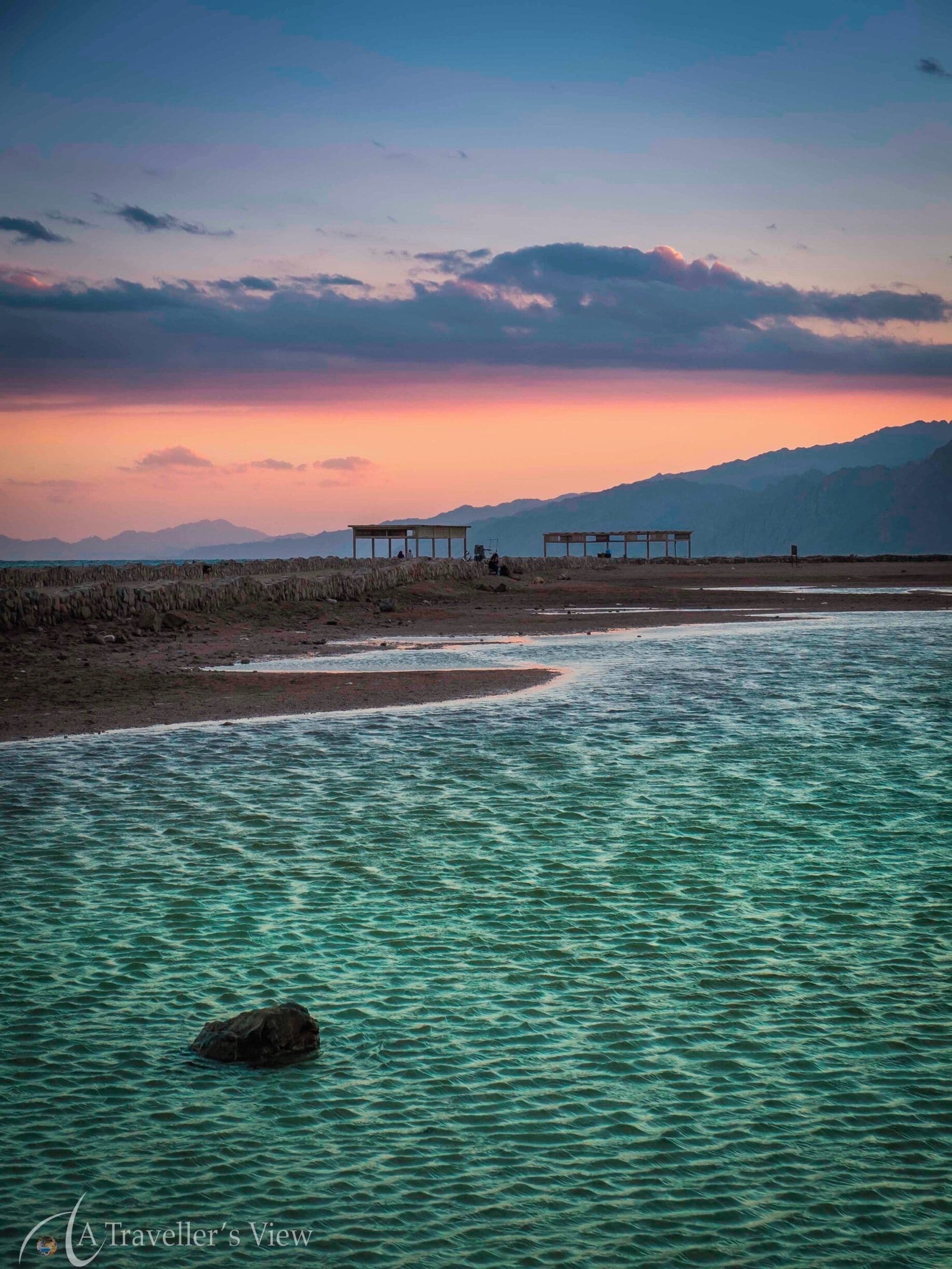 Un río de aguas cristalinas, una roca en primer plano y dos construcciones de madera al otro lado.
