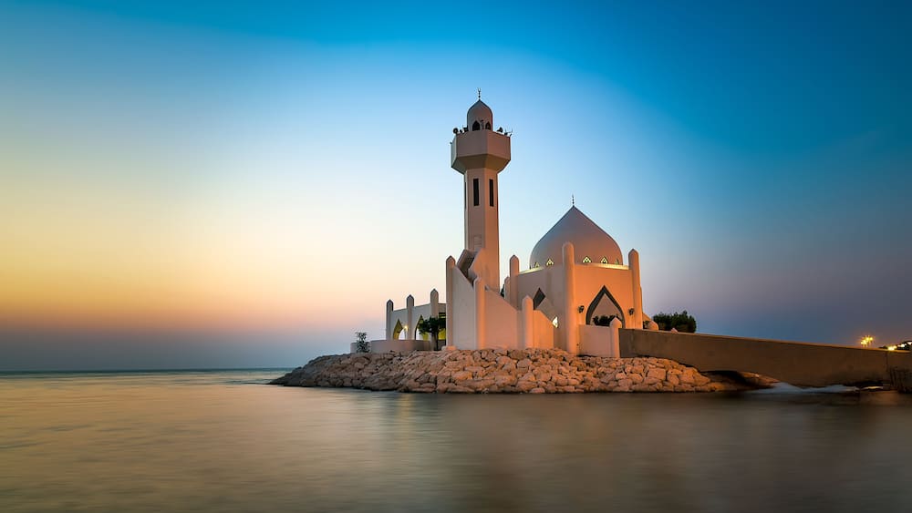 Une mosquée blanche, dotée d’un minaret imposant, se dresse sur une île rocheuse, entourée d’eaux calmes.