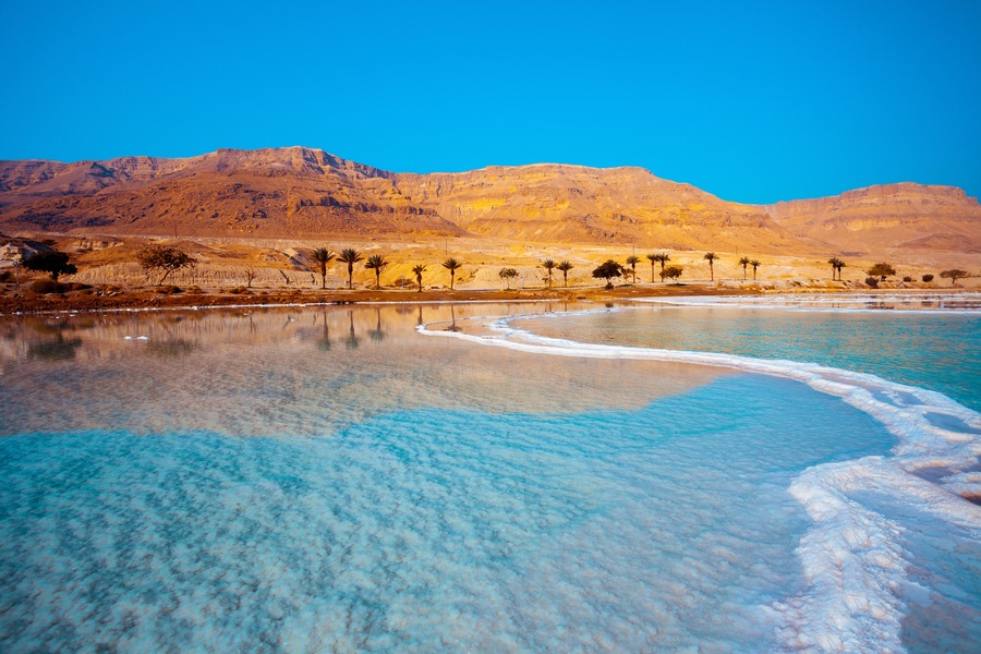 Un lago turquesa y transparente con orilla de arena, palmeras y montañas al fondo.