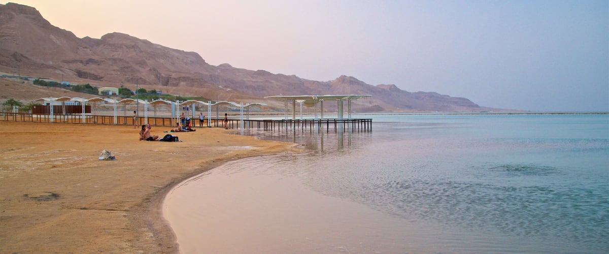 A beach with people sunbathing and a pier extending into calm waters, surrounded by mountains.