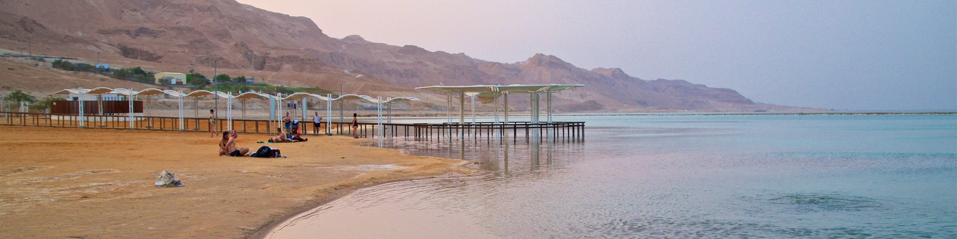 A beach with people sunbathing and a pier extending into calm waters, surrounded by mountains.