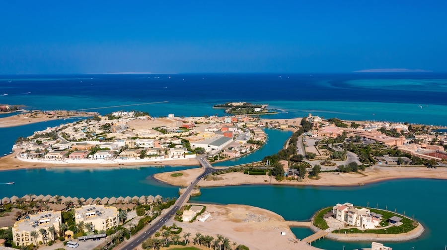 Vista aérea de un complejo turístico frente al mar, con aguas azules cristalinas, una playa de arena y una combinación de edificios residenciales y comerciales.