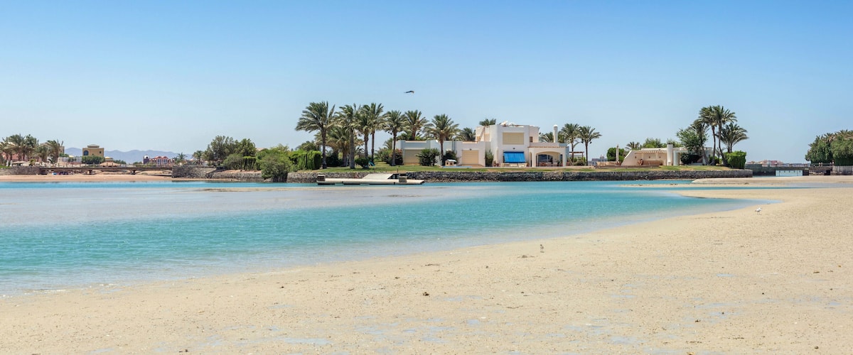 A beach with clear turquoise waters, a sandy shore, and a modern house with a blue roof.