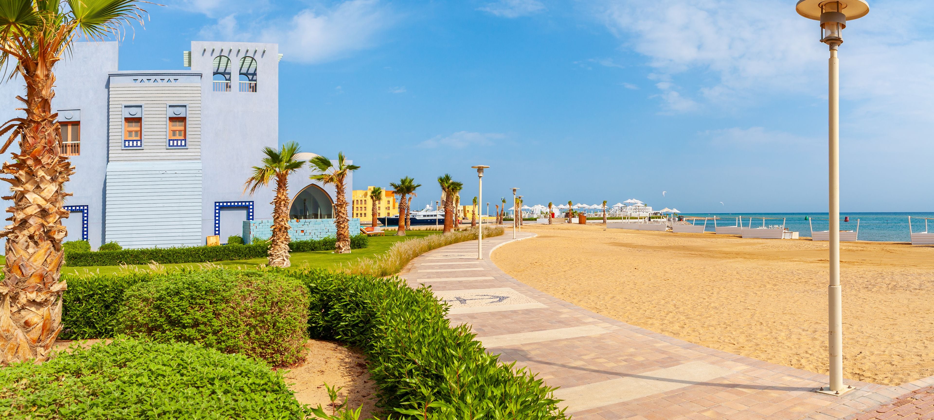 Une promenade en bord de mer bordée de palmiers, une plage et un bâtiment moderne doté de fenêtres en arc de cercle.