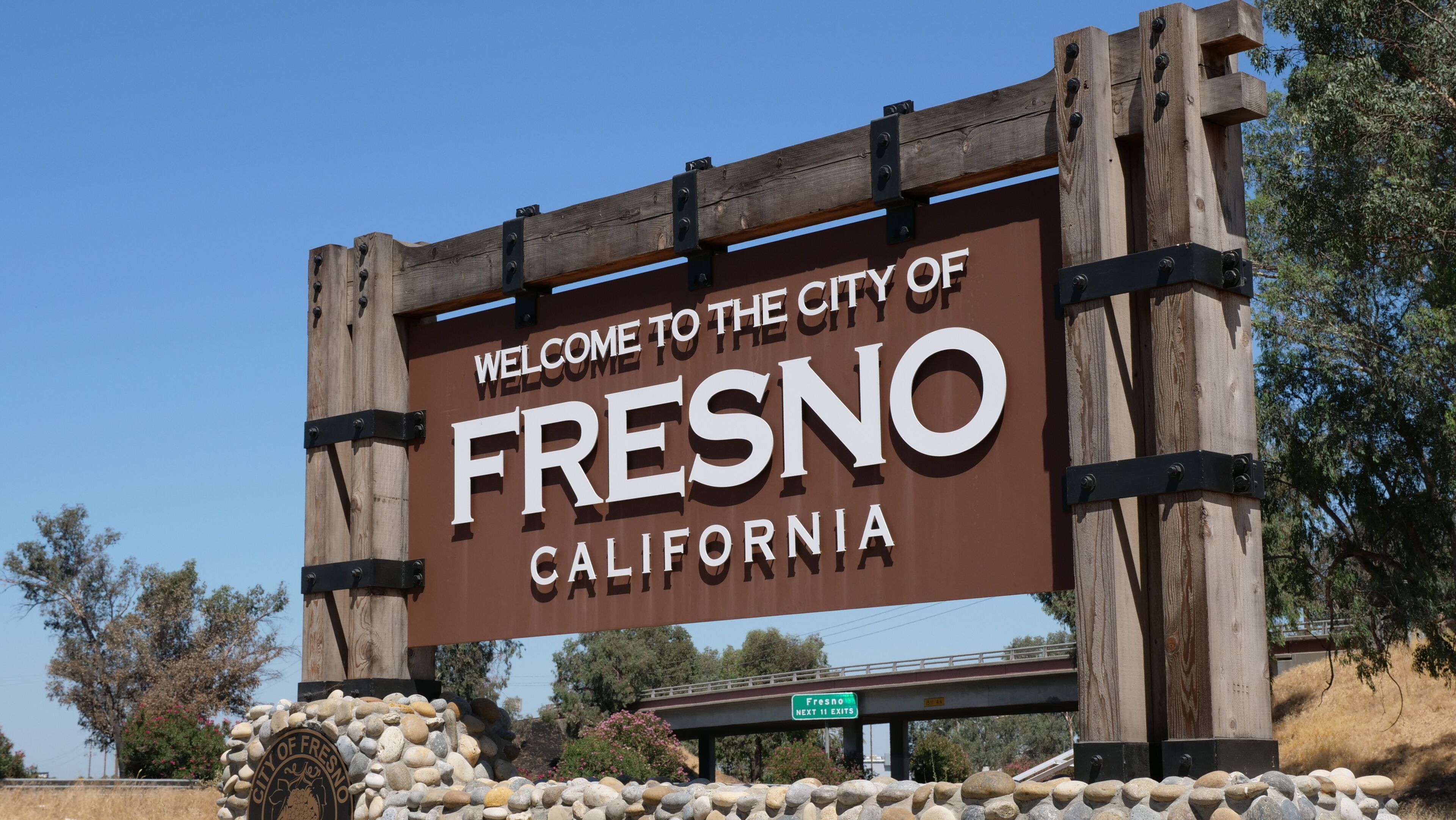 A welcome sign for the City of Fresno, California, with a stone wall and a bridge in the background.