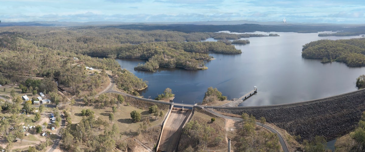 A dam with a road passing over it, surrounded by forested hills and a large lake.
