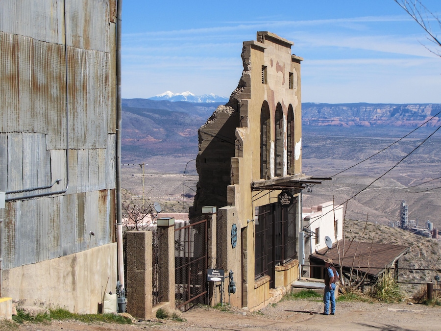 Un bâtiment délabré avec un portail en métal, une personne se tenant à proximité, et un paysage montagneux en arrière-plan.