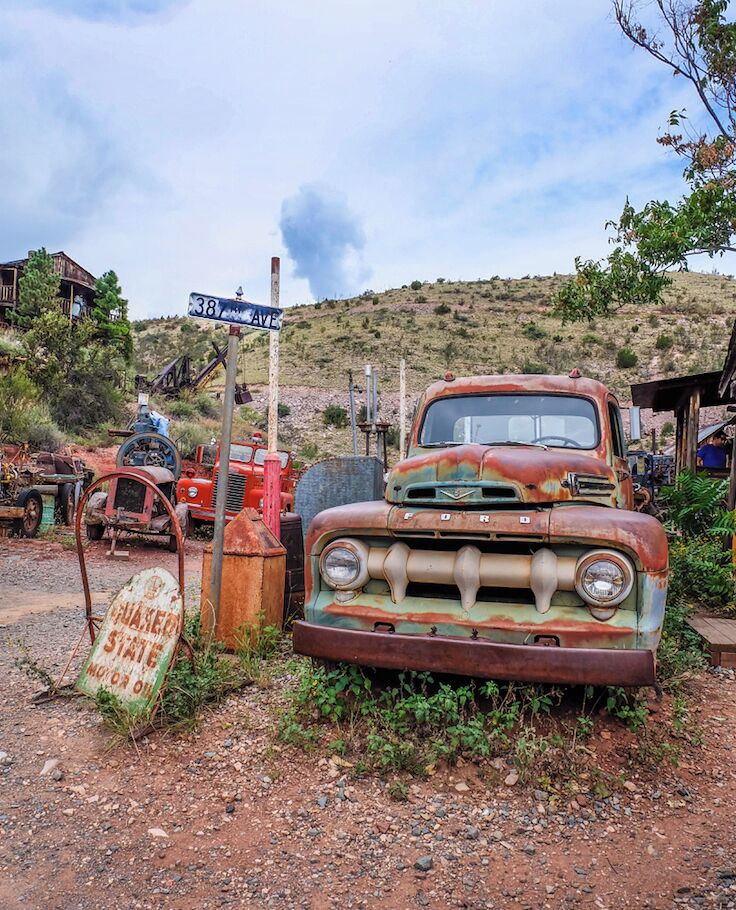 An old, rusted Ford truck on a dirt road with a '38 AVE' sign and a 'CHARGED STATE' sign nearby.