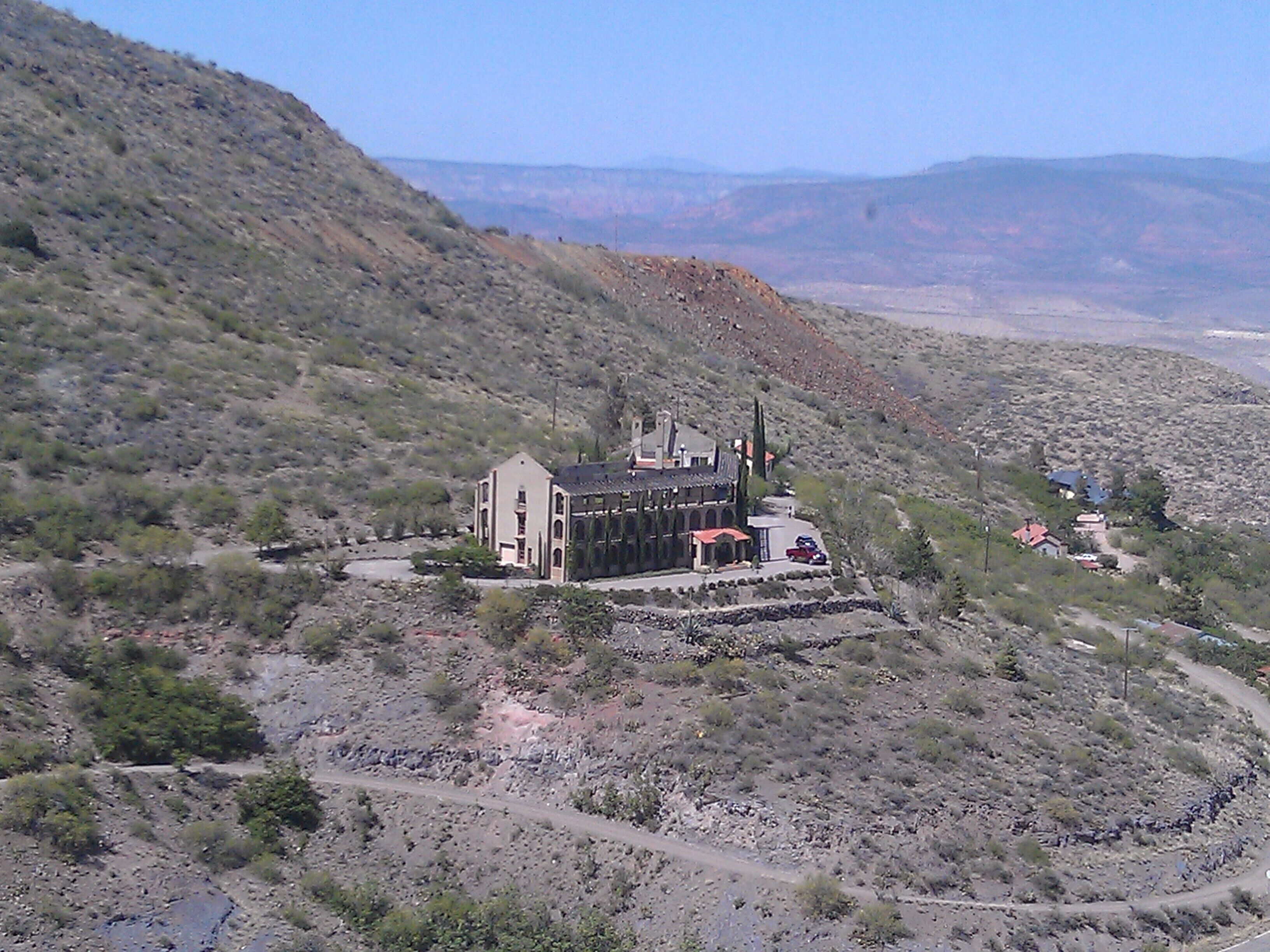 A large, historic building with a red roof and multiple chimneys, surrounded by a rugged, hilly landscape with sparse vegetation.