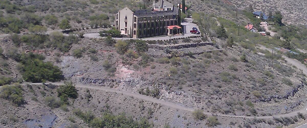 A large, historic building with a red roof and multiple chimneys, surrounded by a rugged, hilly landscape with sparse vegetation.