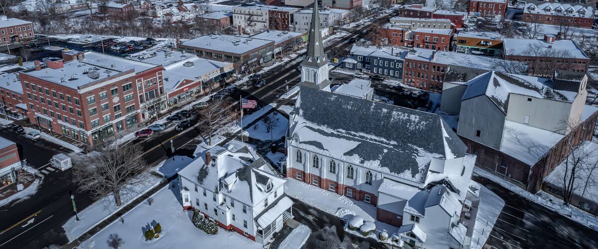 A snow-covered town with a prominent church steeple, red brick buildings, and a clear blue sky.