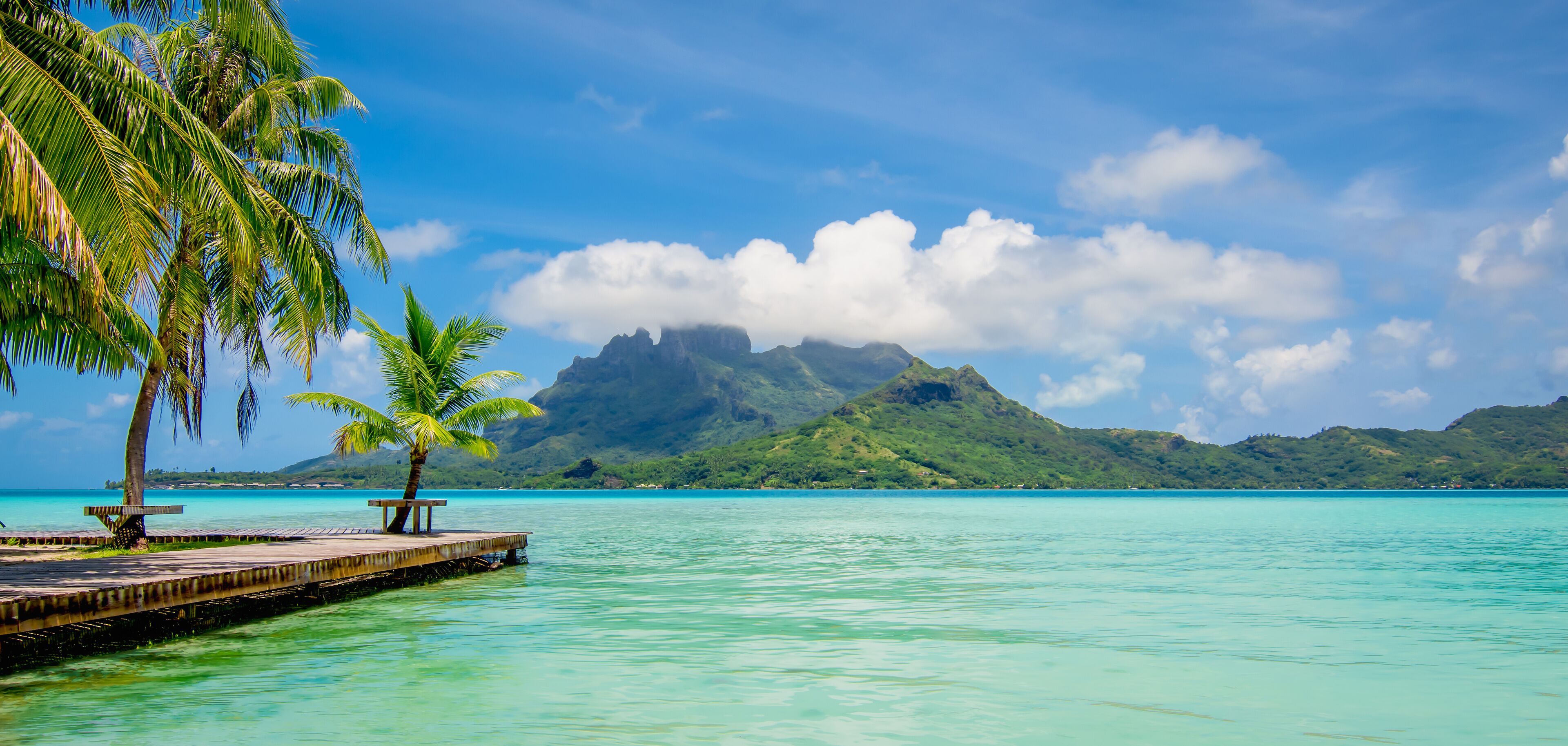 A tropical beach with clear turquoise water, palm trees, a wooden pier, and a mountain in the background.