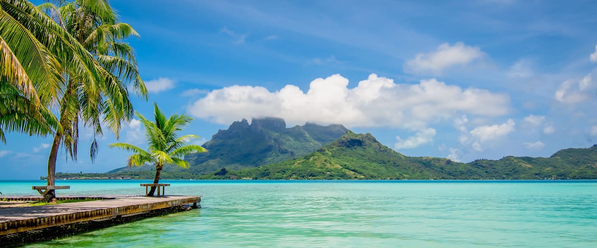 A tropical beach with clear turquoise water, palm trees, a wooden pier, and a mountain in the background.