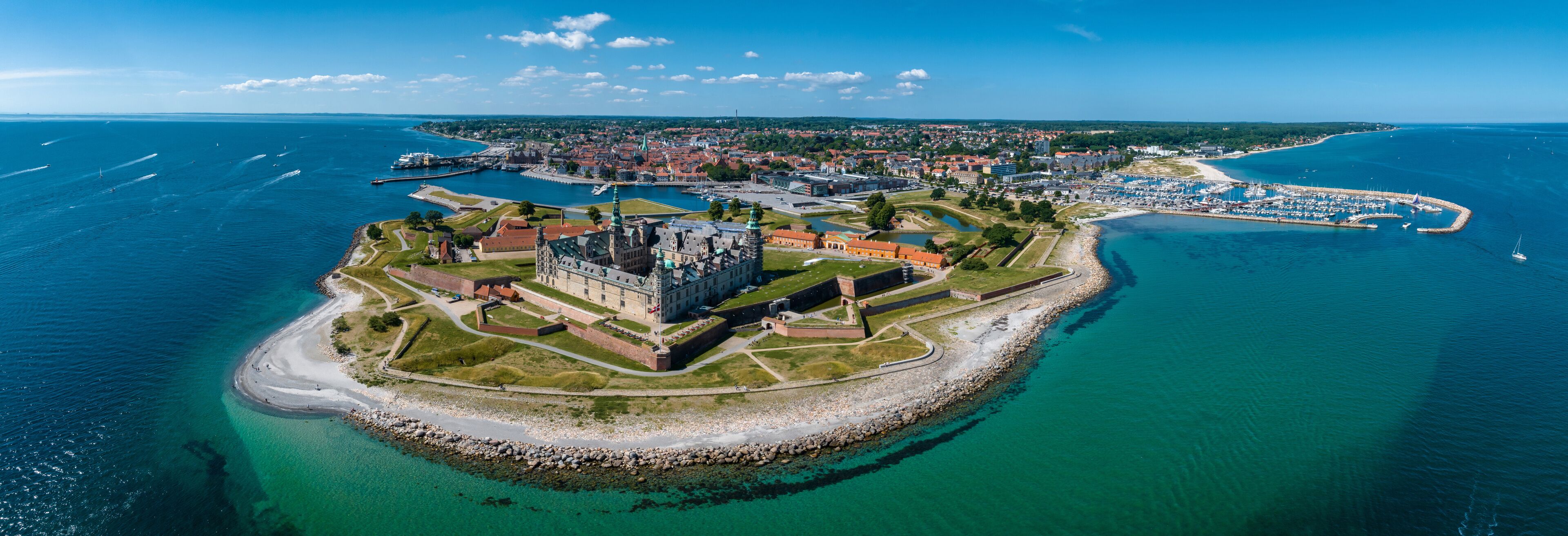 An aerial view of a coastal town with a prominent castle, a marina, and a beach.