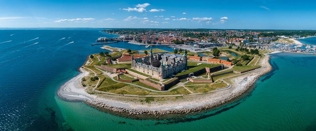 An aerial view of a coastal town with a prominent castle, a marina, and a beach.