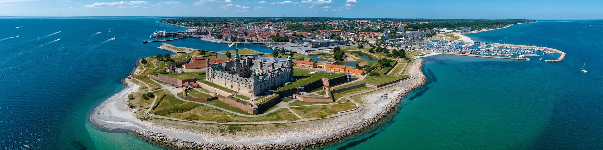 An aerial view of a coastal town with a prominent castle, a marina, and a beach.