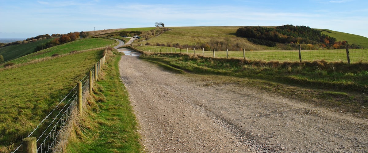 A rural dirt road winding through green fields with a fence on one side and a hill in the distance.