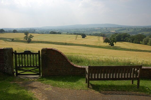 A rural landscape with a brick wall, a wooden gate, and a bench overlooking a field.