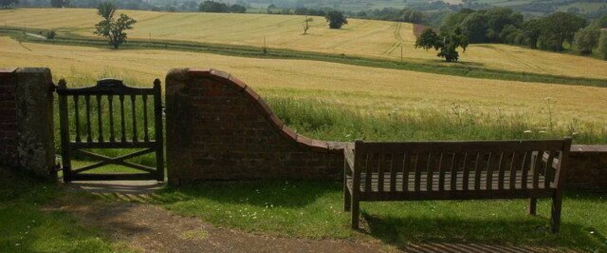 A rural landscape with a brick wall, a wooden gate, and a bench overlooking a field.