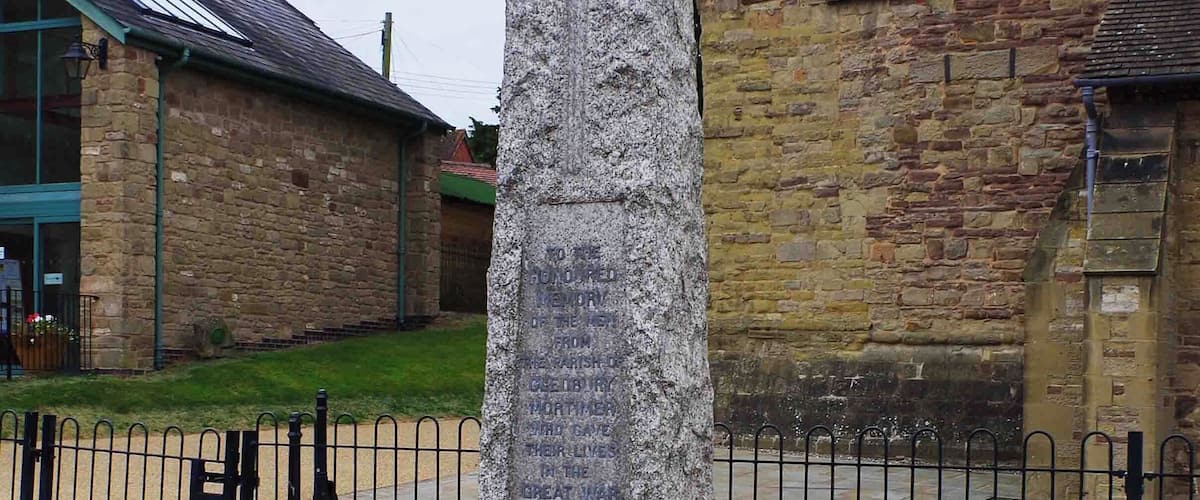A stone cross monument with engraved names, located in a courtyard with a stone building in the background.