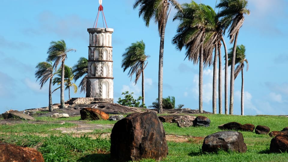 A lighthouse on a grassy hill with palm trees and large rocks.