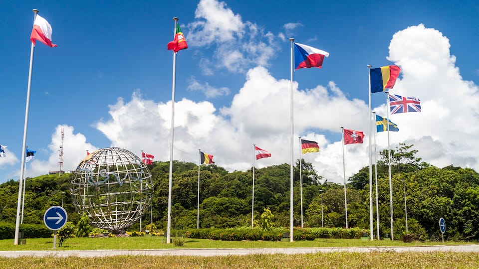 A globe surrounded by various national flags, a blue directional sign, and lush greenery under a clear blue sky.
