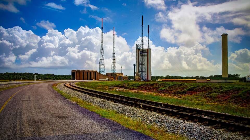 A railway track curving to the right with a launch tower and a tall chimney in the background.