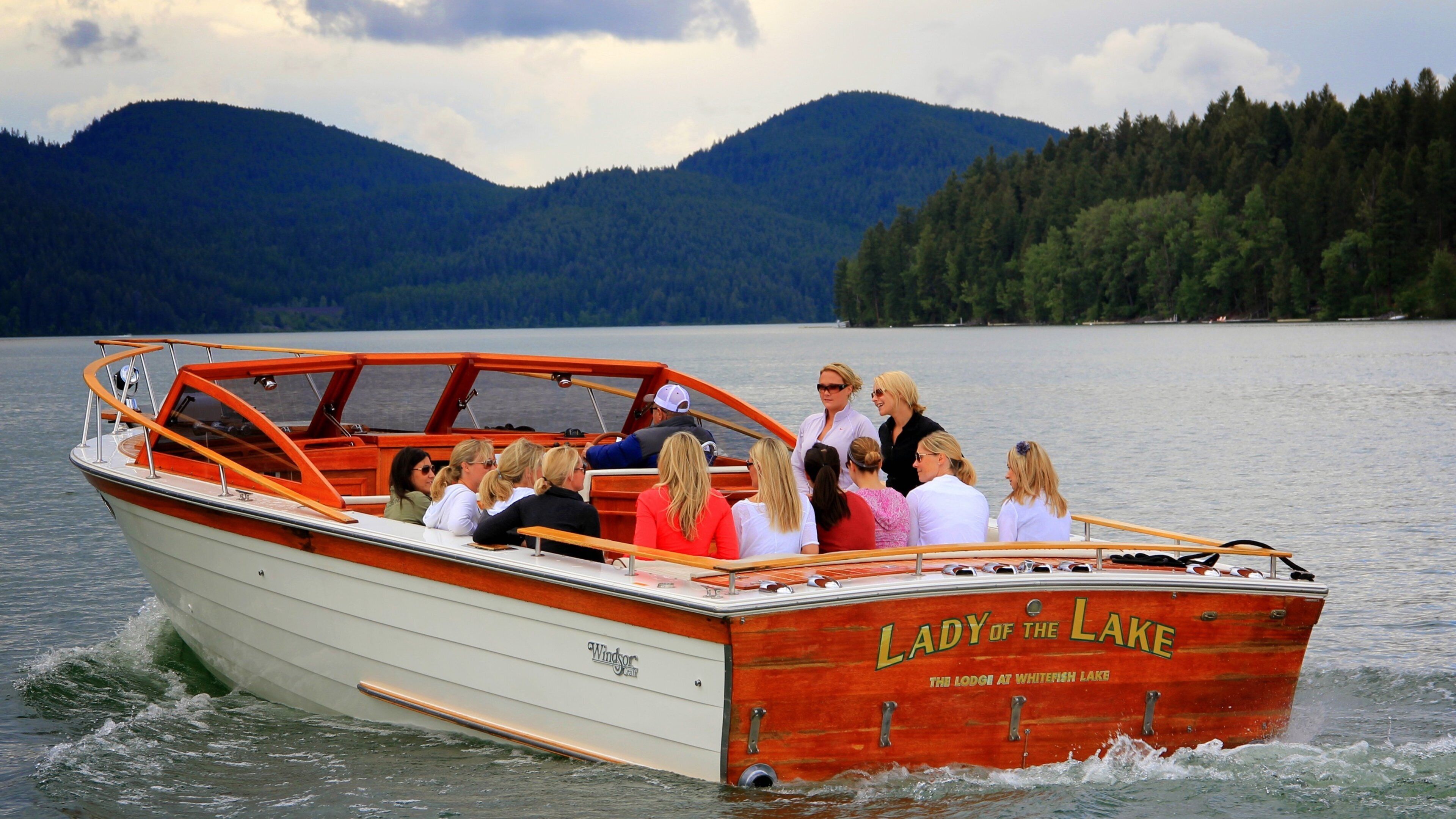 A boat named "Lady of the Lake" with people on board, moving on a lake with mountains in the background.