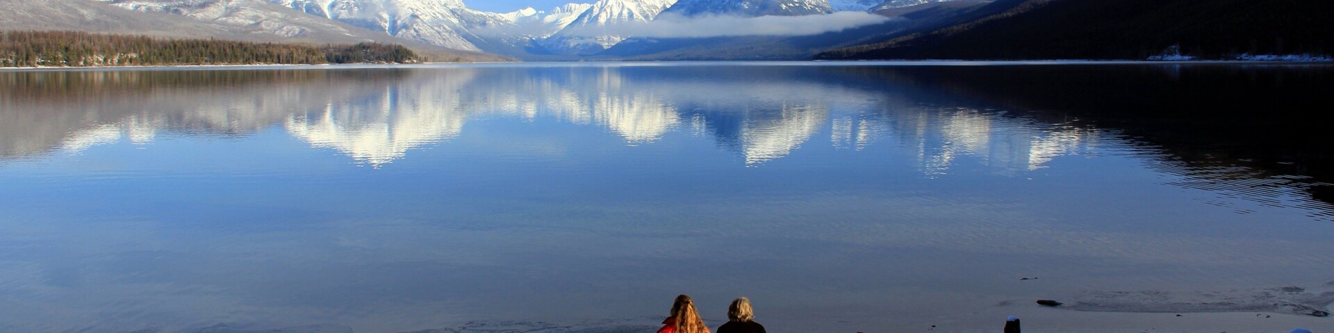 Una familia de pie en una orilla cubierta de nieve, con un lago tranquilo y montañas nevadas al fondo.