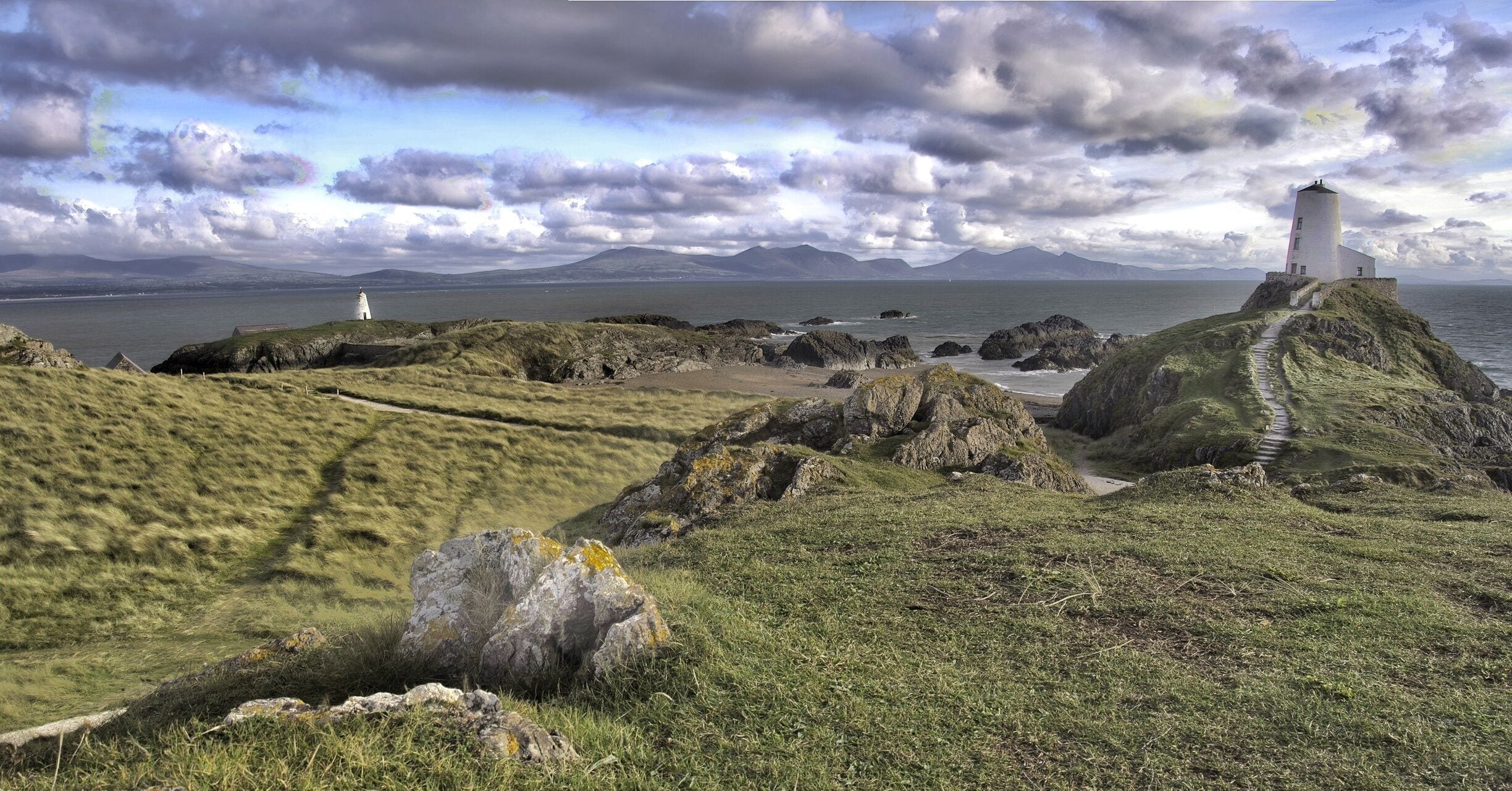 A coastal landscape with a lighthouse, rocky terrain, grassy hills, and a distant mountain range.
