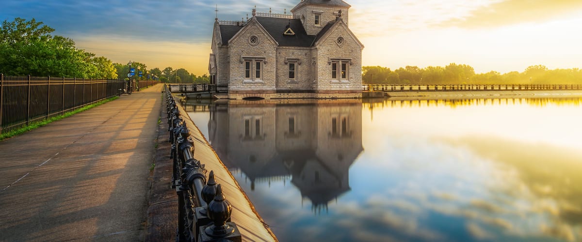 A historic stone building with a reflection in calm water, surrounded by a fence and trees.